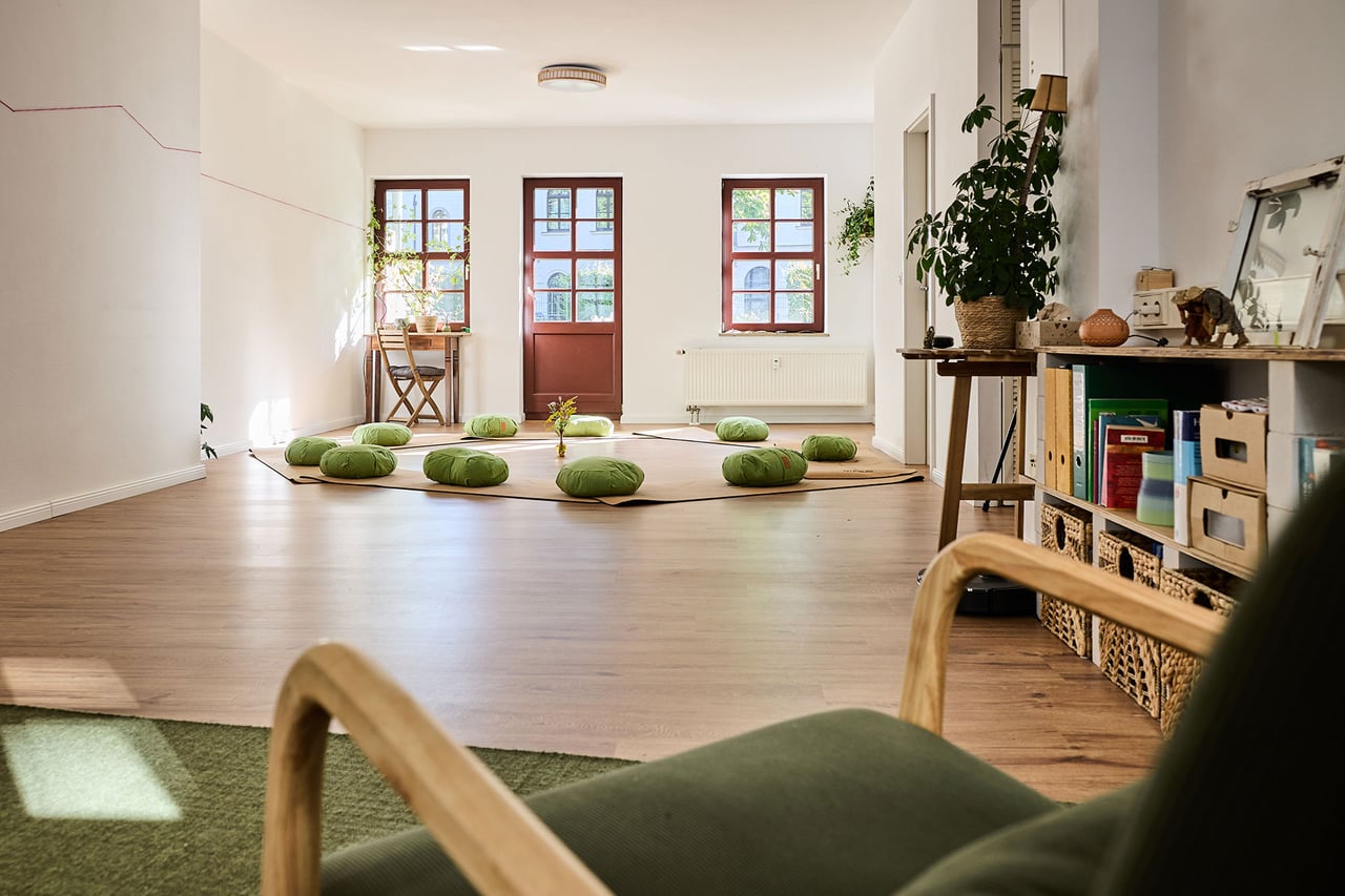 Blick durch den Praxisraum auf die Meditationskissen, mit Sessel und Regal im Vordergrund